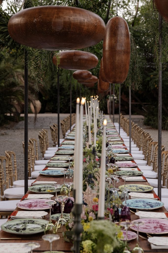 Tavolo da pranzo all'aperto con decorazioni floreali, candele e grandi sculture di legno sospese, perfetto per matrimoni e eventi eleganti, ambientazione naturale e raffinata.