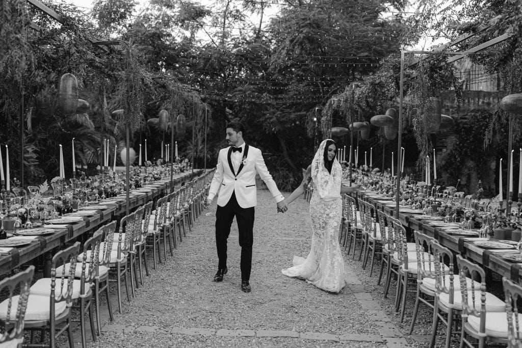 Un uomo e una donna in abiti da sposa e sposo si tengono per mano in un elegante allestimento di matrimonio all'aperto, con tavoli decorati e luci soffuse, pronti per un evento speciale.