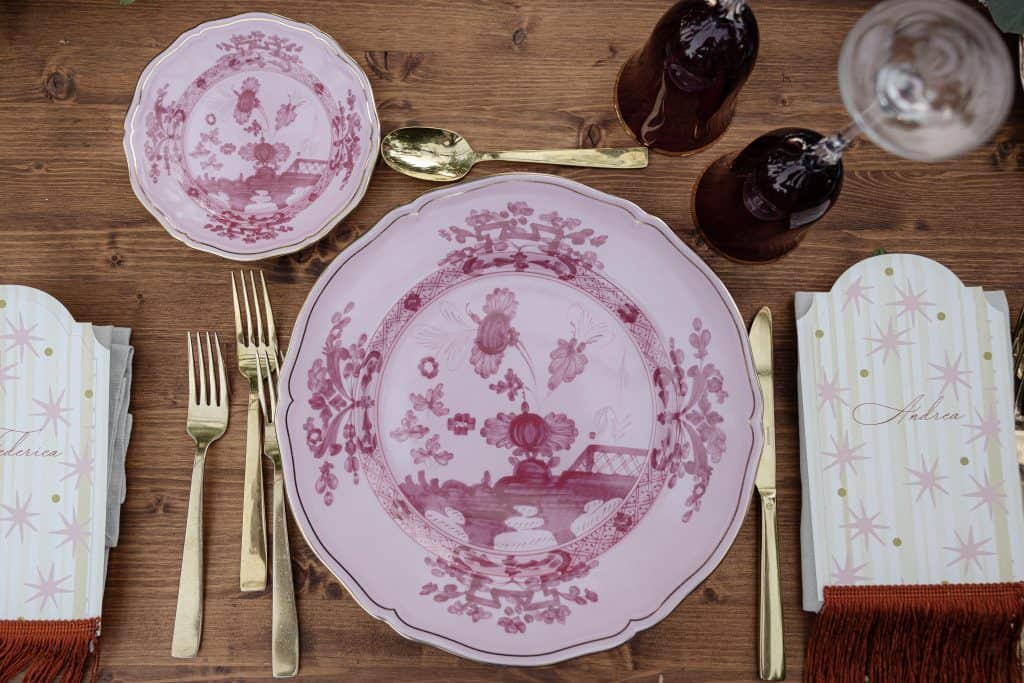 Piatti eleganti in porcellana rosa con motivi floreali e paesaggistici, perfetti per matrimoni e eventi speciali, ambientati su un tavolo in legno rustico.