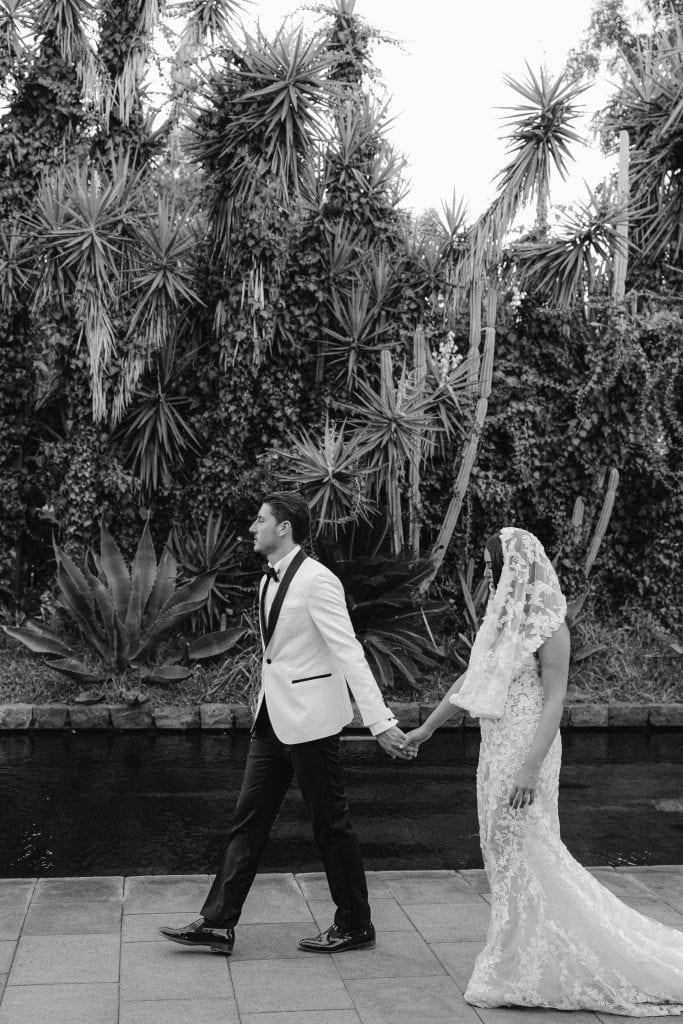 Un uomo e una donna in abiti da sposa e sposo si tengono per mano davanti a una vegetazione tropicale, in un matrimonio elegante e romantico.