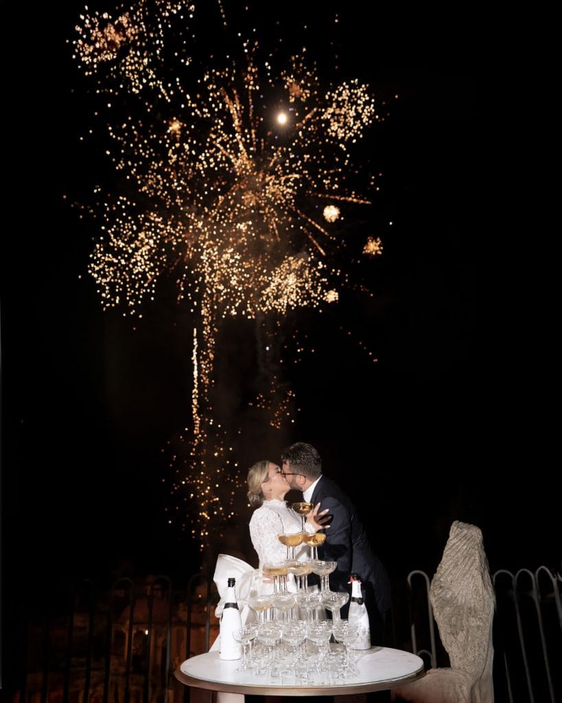 Festa di matrimonio con fuochi d'artificio, coppia che si bacia, brindisi con champagne, allestimento elegante, ChiaraB Events Wedding Planner, evento romantico e memorabile.