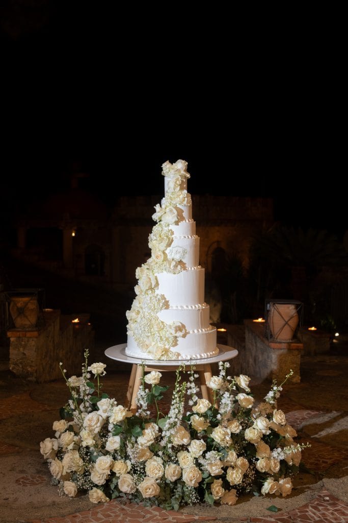 Torta nuziale elegante decorata con fiori bianchi, perfetta per matrimoni raffinati, su un tavolo decorato con un'ampia composizione floreale, ambientazione romantica e sofisticata.