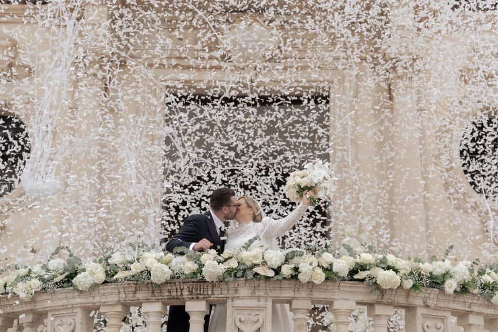 Elegante coppia di sposi che si bacia sul balcone decorato con fiori bianchi, durante il matrimonio organizzato da ChiaraB Events Wedding Planner.