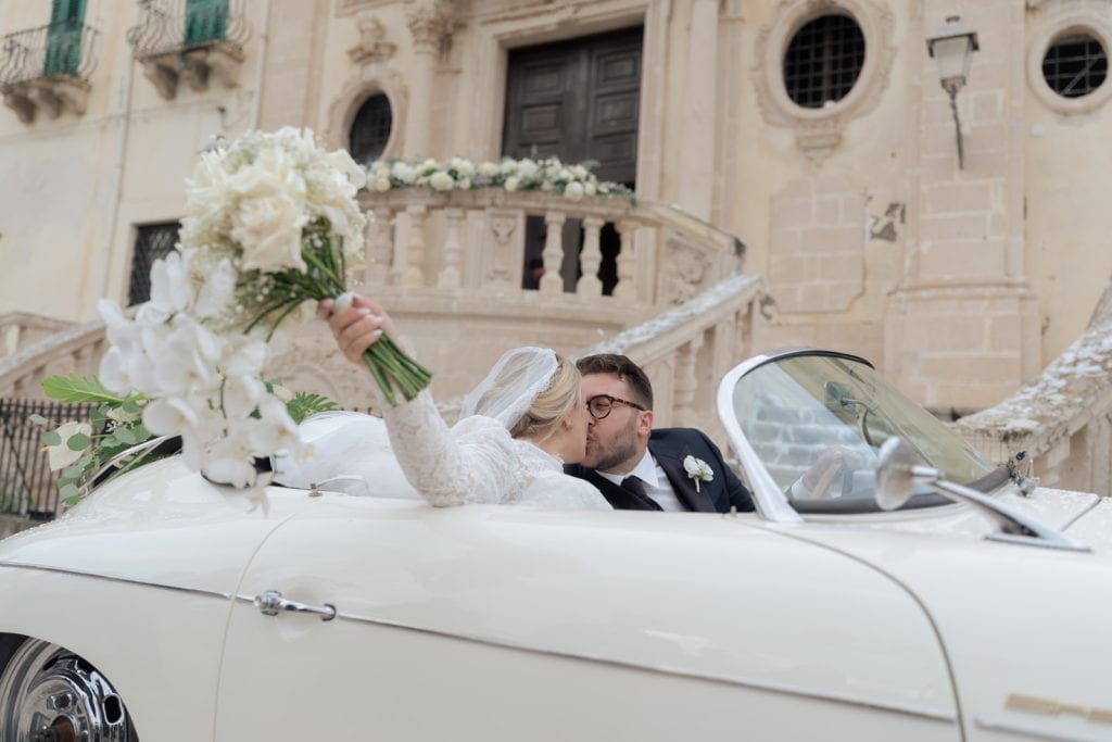 Coppia di sposi in auto d'epoca durante il matrimonio, con sfondo storico e architettura elegante, perfetta per servizi di wedding planning e cerimonie romantiche in Italia.