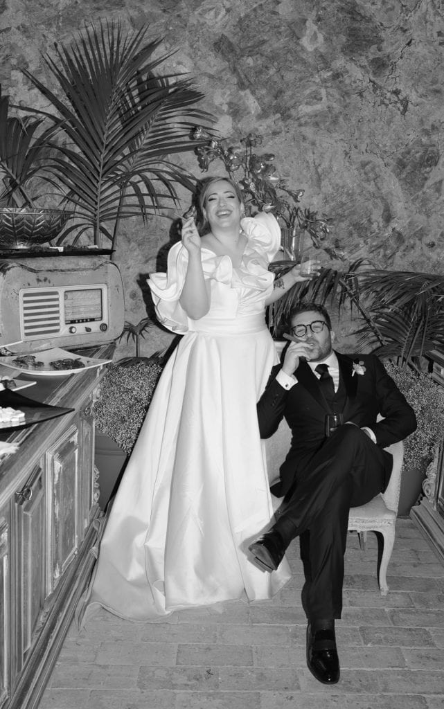 Elegante coppia di sposi in un matrimonio, con la sposa in abito lungo e il sposo in completo nero, ambientazione raffinata con piante tropicali e decorazioni vintage.