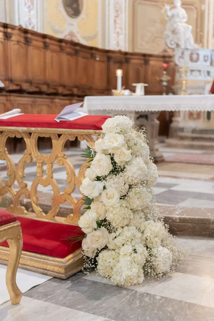 Un bouquet di fiori bianchi decorativo e elegante, posizionato accanto a un banco con dettagli dorati e un cuscino rosso, in un ambiente di chiesa o location per matrimoni.