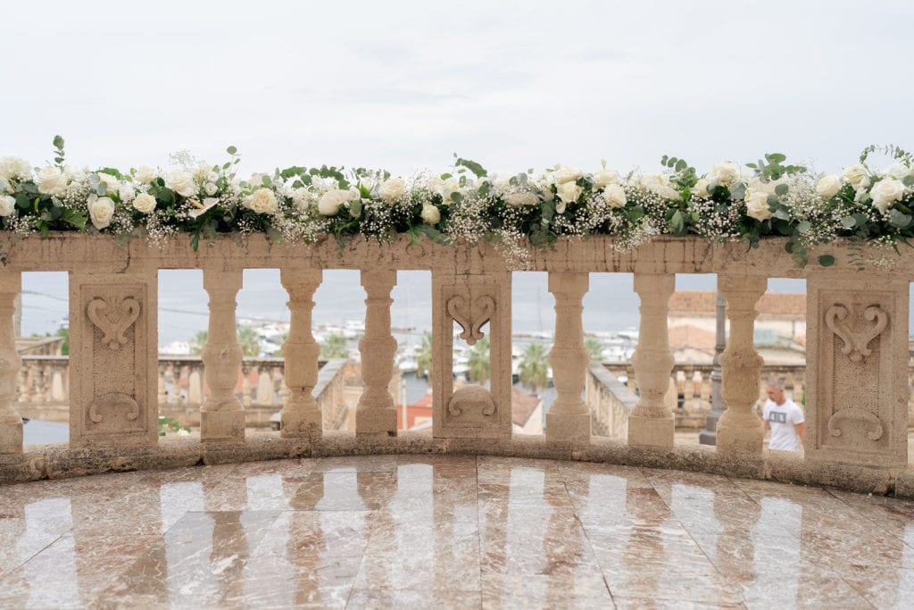 Decorazione floreale elegante su balaustra in pietra per matrimonio, vista panoramica sulla città, perfetta per wedding planner e eventi romantici all'aperto.