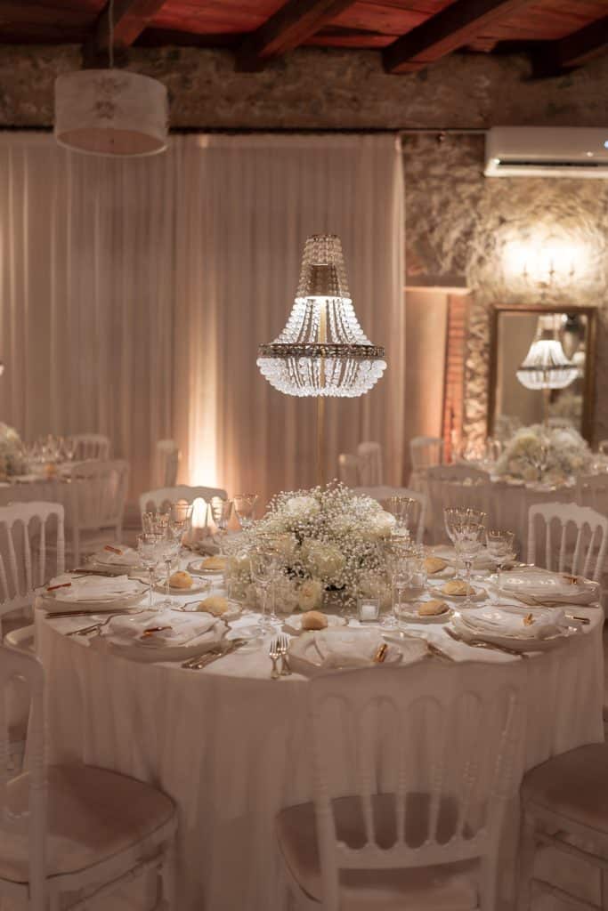 Elegante tavola nuziale decorata con fiori bianchi e candele, ambientata in un locale rustico con pareti in pietra e soffitto in legno, perfetta per matrimoni raffinati e personalizzati.