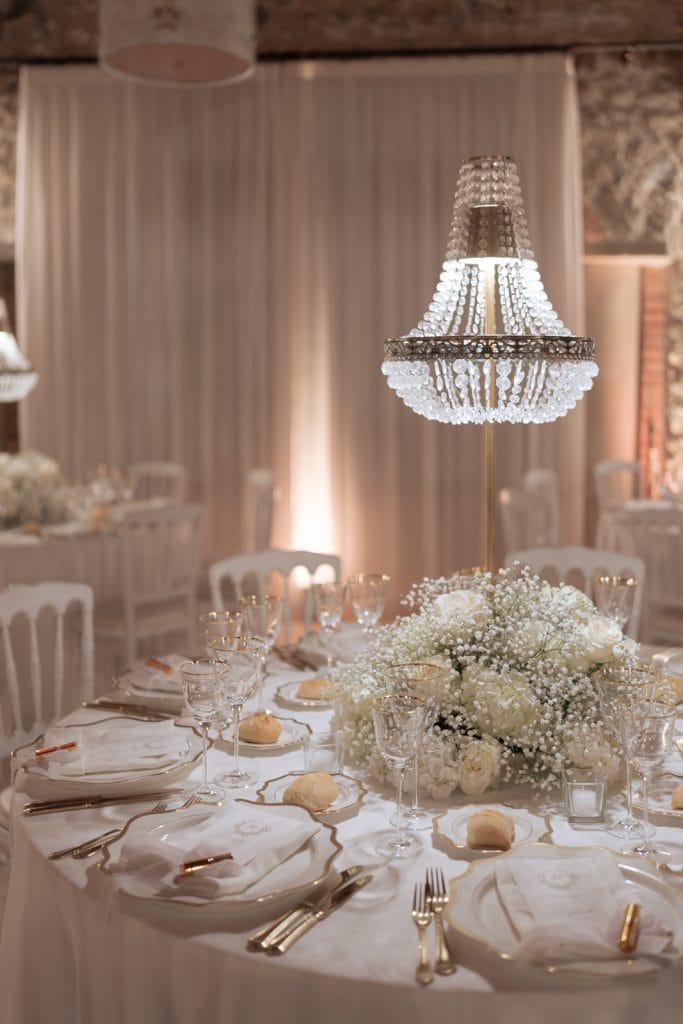 Elegante tavola da matrimonio decorata con fiori bianchi e cristalli, illuminata da un lampadario di cristallo, perfetta per eventi di lusso e matrimoni romantici.