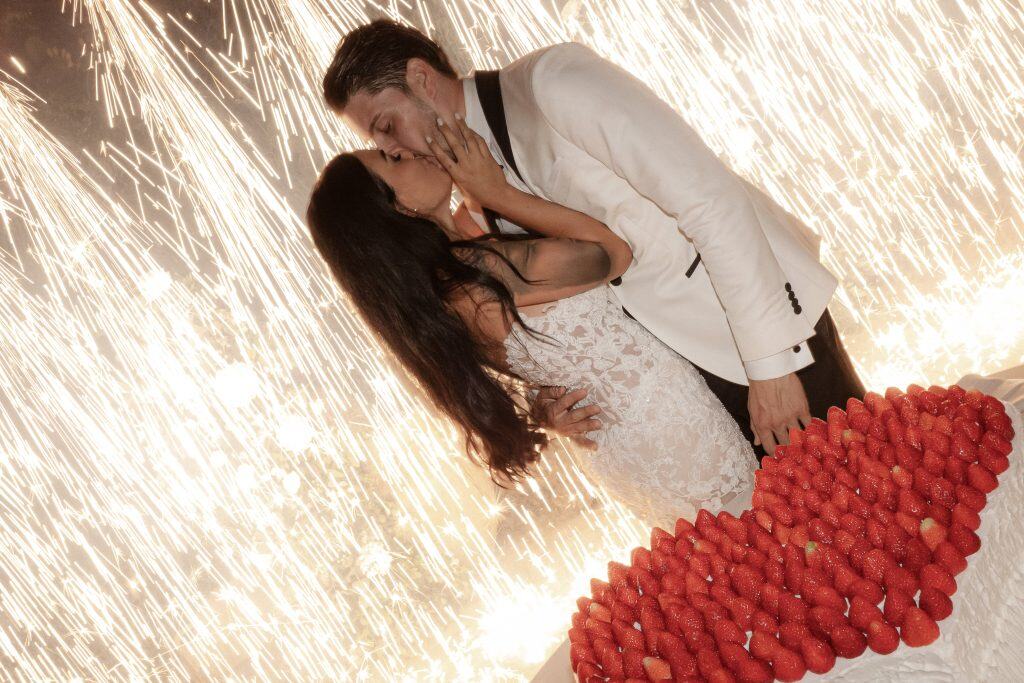 Un bacio romantico tra una coppia di sposi con fuochi d'artificio sullo sfondo, simbolo di amore e celebrazione del matrimonio, con un cuore di fragole sul tavolo. Perfetto per servizi di wedding plan.