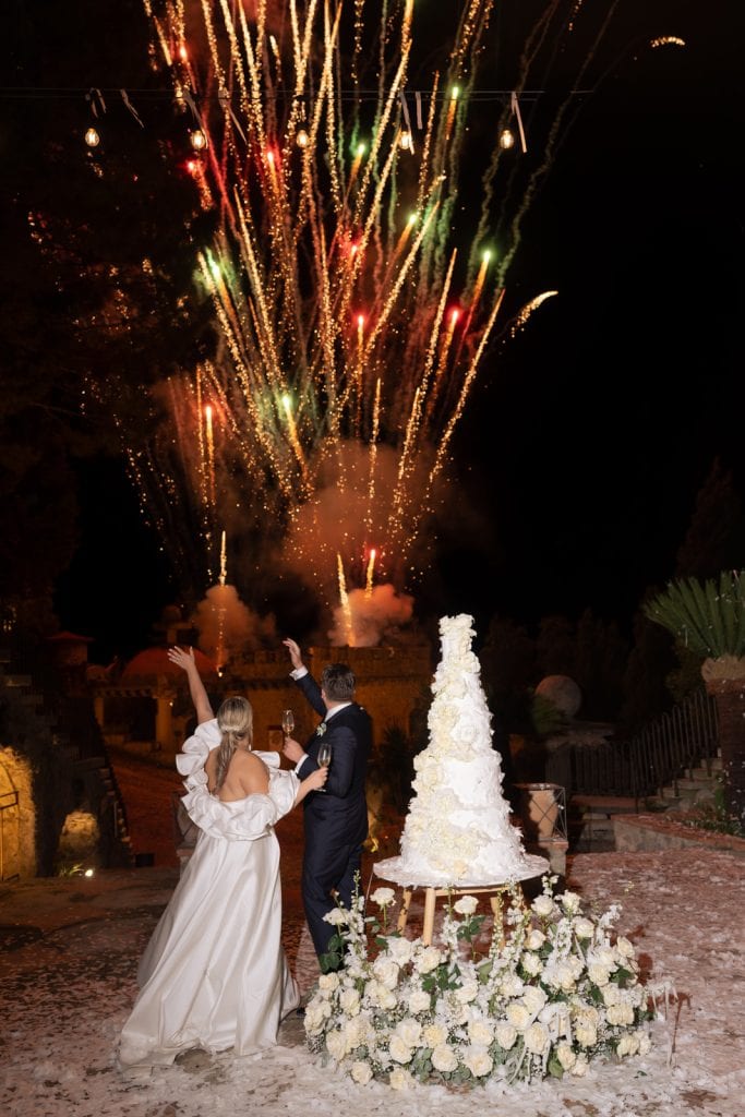 Un fuochi d'artificio esplodono nel cielo notturno durante un matrimonio, con una coppia di sposi che si tiene per mano davanti a una torta nuziale decorata con fiori bianchi, in un'atmosfera magica e.