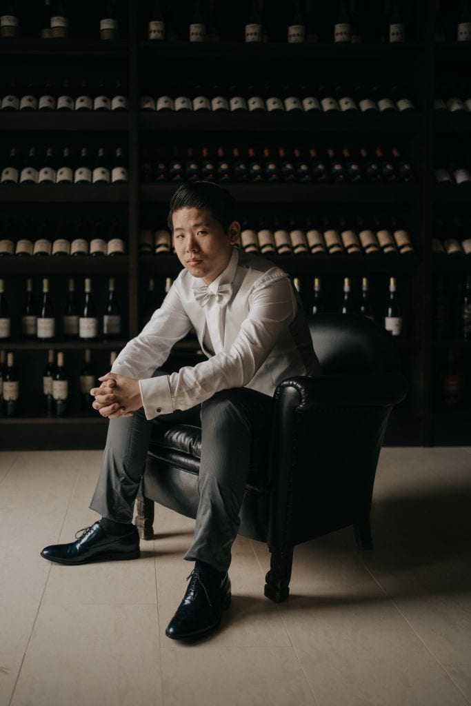 Elegante uomo seduto su poltrona in un ambiente con scaffali di bottiglie di vino, rappresentando un professionista del wedding planning e degli eventi di alta qualità.
