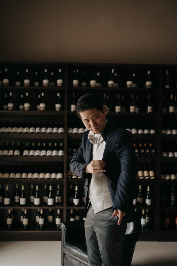Elegante uomo in abito da sera in un ambiente raffinato con scaffali di bottiglie di vino sullo sfondo, perfetto per eventi e matrimoni di lusso.