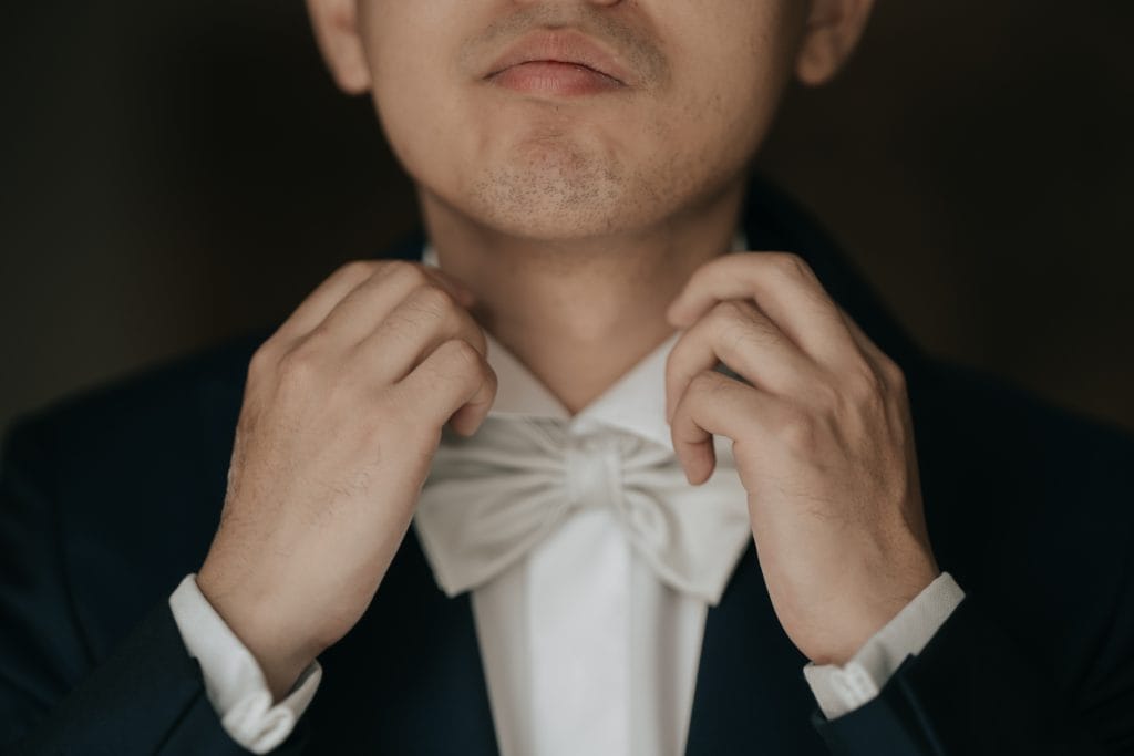 Elegante uomo in abito da sposo con papillon bianco, pronto per il matrimonio, in un ambiente sobrio e raffinato. Perfetto per servizi fotografici di wedding planning e cerimonie di lusso.