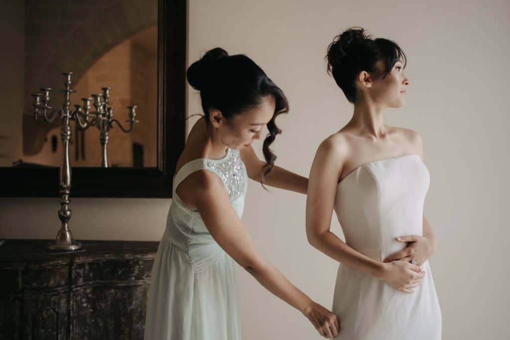 Un'addetta alle nozze aiuta una sposa a prepararsi, indossando un abito da sposa elegante in un ambiente raffinato. Servizi di wedding planning professionale e personalizzato per matrimoni indimentica.