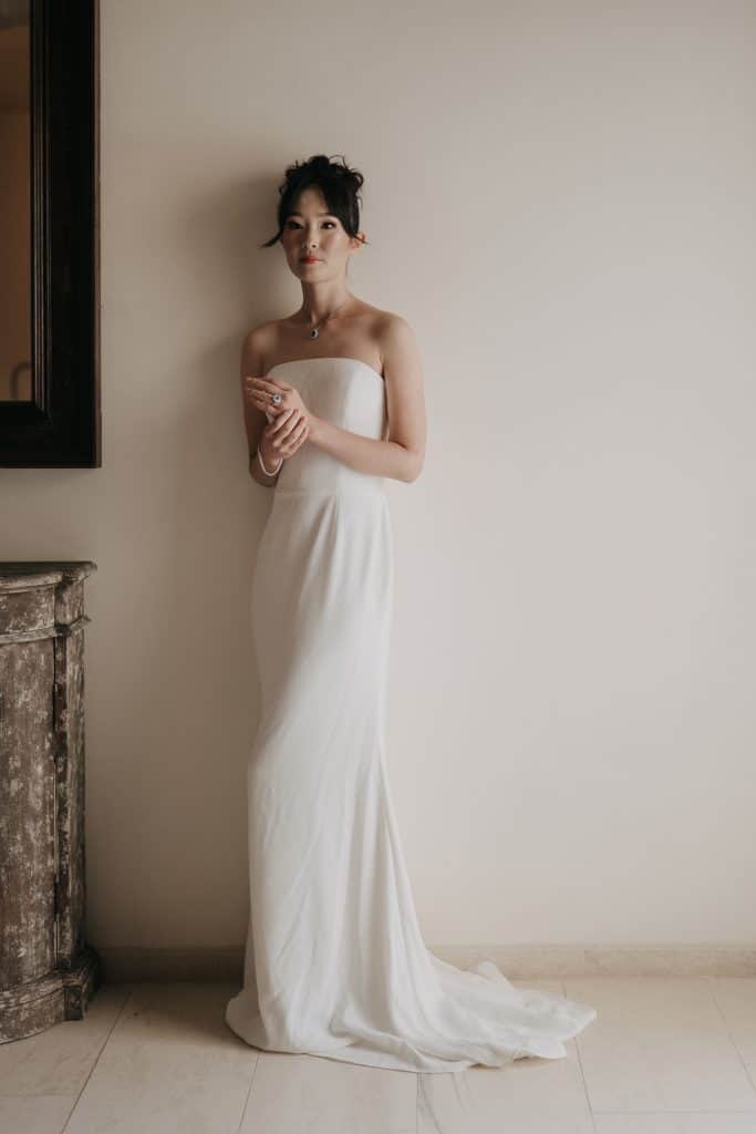 Elegante sposa in abito bianco lungo, pronta per il matrimonio, in un ambiente minimalista con parete neutra e arredi classici. Perfetta per servizi fotografici di wedding planning e cerimonie di luss.