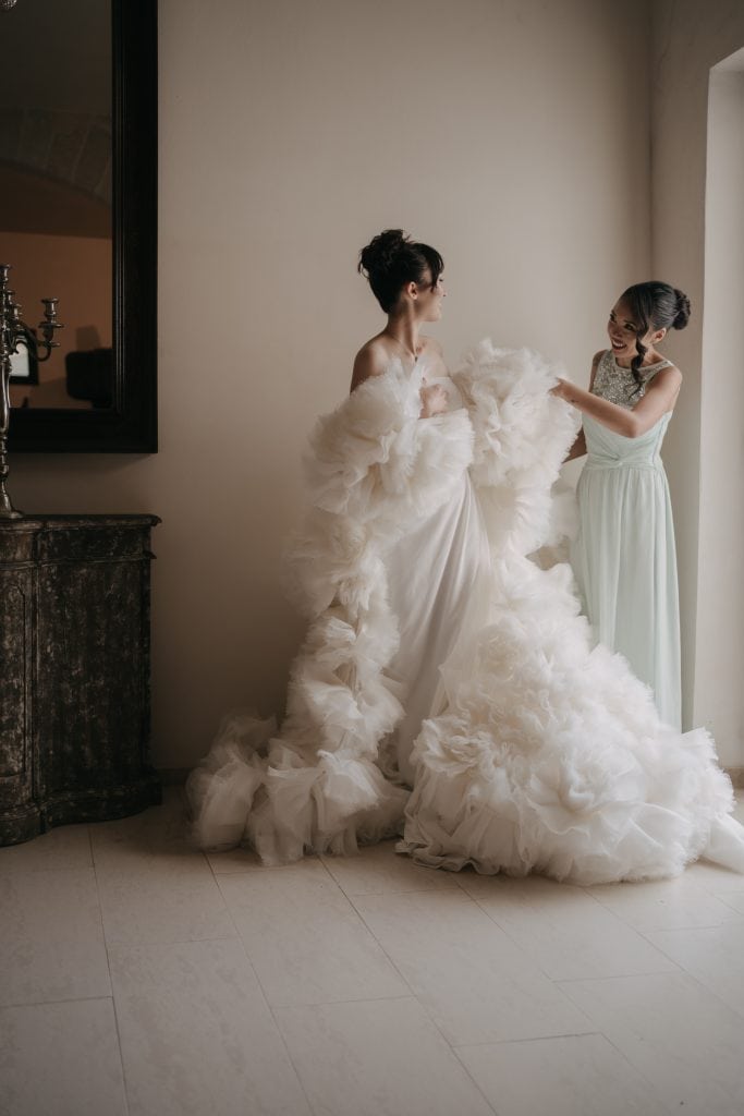Lunghezza ALT tag: Donna in abito da sposa con ampio strascico e acconciatura raccolta, assistita da una donna in abito lungo, in un ambiente elegante e luminoso, perfetto per servizi di wedding plann.