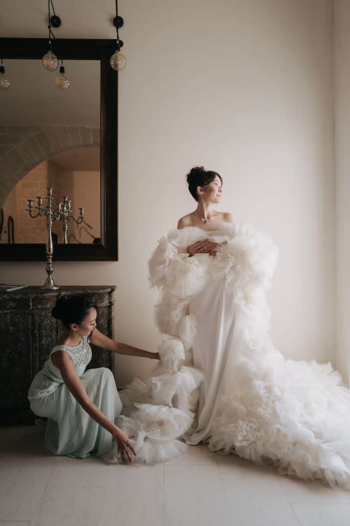Elegante sposa in abito da cerimonia con dettagli in pizzo e tulle, assistita da una damigella in un ambiente raffinato e luminoso, perfetto per servizi di wedding planning e organizzazione matrimoni.