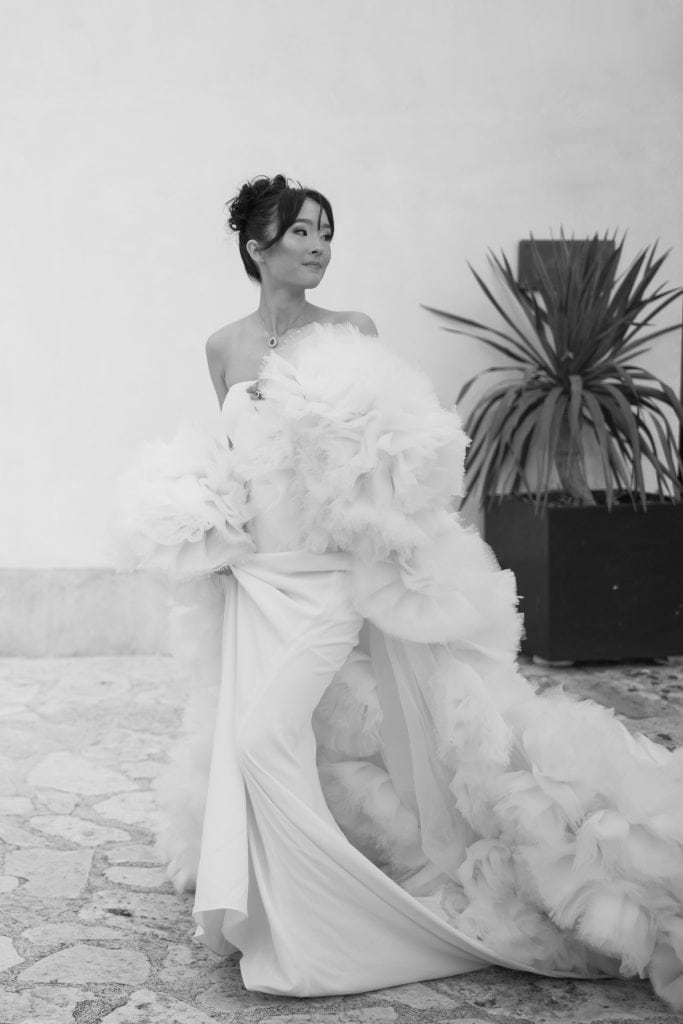 Un'illustrazione di una donna in abito da sposa elegante, con dettagli di pizzo e tessuti voluminosi, in un ambiente minimalista con pianta decorativa sullo sfondo. Perfetta per servizi di wedding pla.