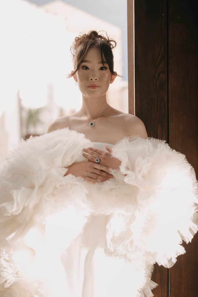 Elegante sposa con abito da matrimonio bianco e dettagli in pelliccia, posa davanti a una porta in legno, pronta per il suo giorno speciale, assistita da ChiaraB Events Wedding Planner.
