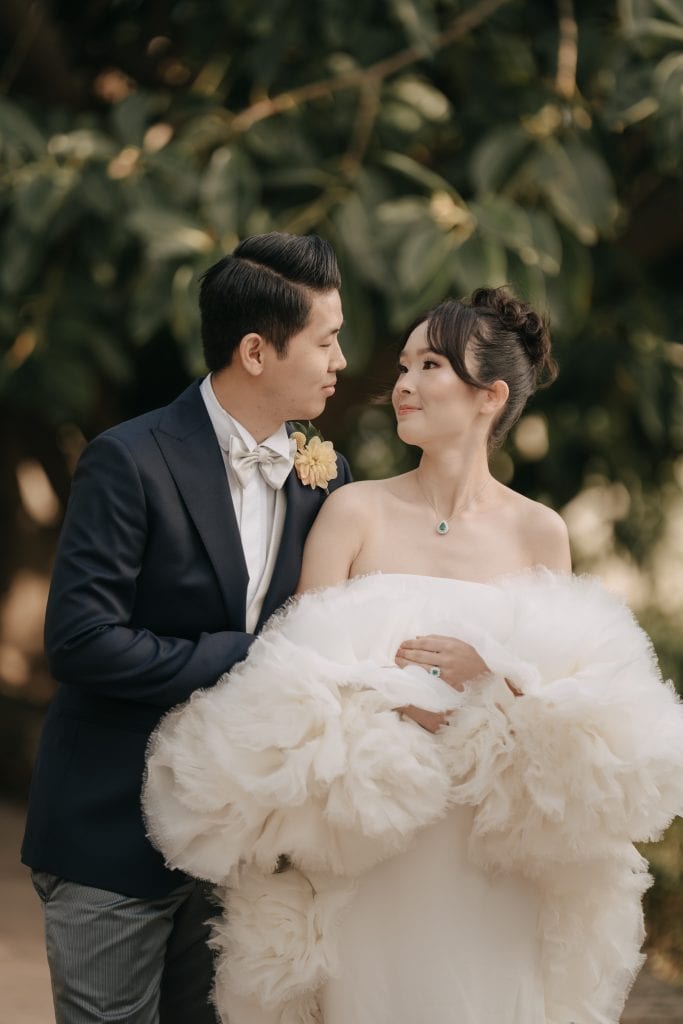 Elegante coppia di sposi in abiti da matrimonio, ambientazione naturale con foglie verdi sullo sfondo, perfetta per servizi di wedding planning e organizzazione eventi nuziali.