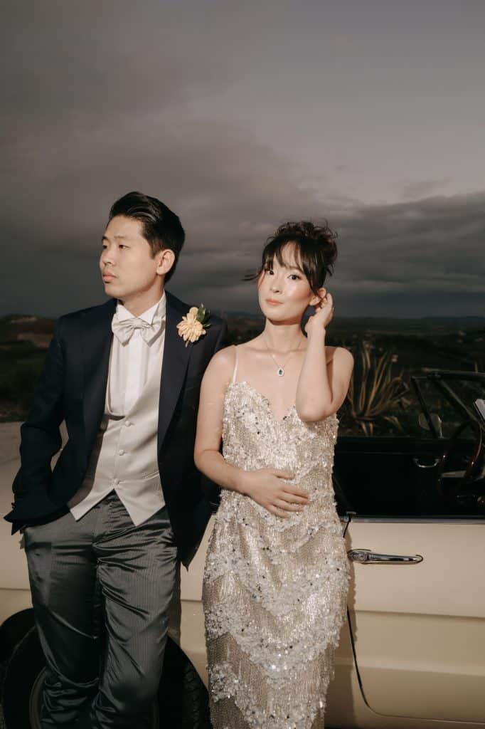 Elegante coppia di sposi in abiti da matrimonio sofisticati, con sfondo di cielo nuvoloso e auto d'epoca, perfetta per servizi di wedding planning e organizzazione eventi di lusso.