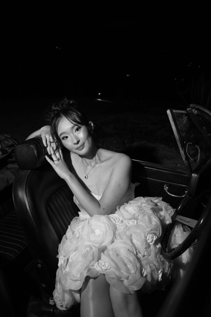 Elegante donna in abito da sposa con dettagli floreali, seduta in auto d'epoca, in un'immagine nera e bianca, rappresentando un servizio di wedding planning di ChiaraB Events, specializzato in matrimo.