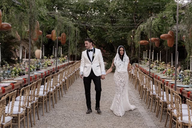 Menyasszony és vőlegény esküvői ruhában, kéz a kézben, szabadtéri esküvőre díszített asztalok között sétálva, romantikus hangulattal és elegáns részletekkel.