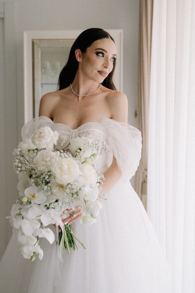 Donna in abito da sposa con bouquet di fiori bianchi, pronta per il matrimonio, ambientazione elegante e luminosa.