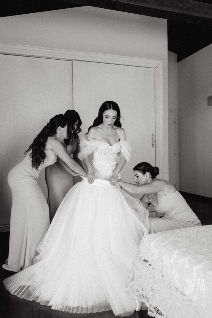 Donne che aiutano la sposa a indossare il vestito da sposa in una stanza moderna, momento di preparazione prima del matrimonio.