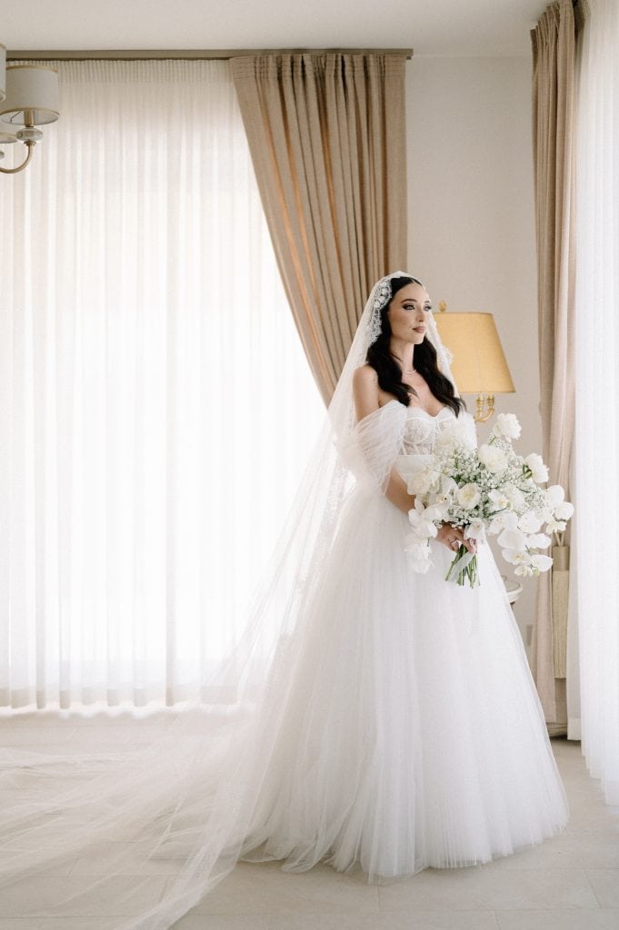 Sposa in abito bianco con velo, in uno studio luminoso con tende beige, pronta per il matrimonio.
