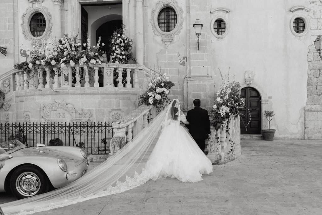 Sposi Alberto e Valeria davanti a una storica chiesa, con abito da sposa con lungo velo e abito da sposo classico, decorazioni floreali e auto d'epoca.