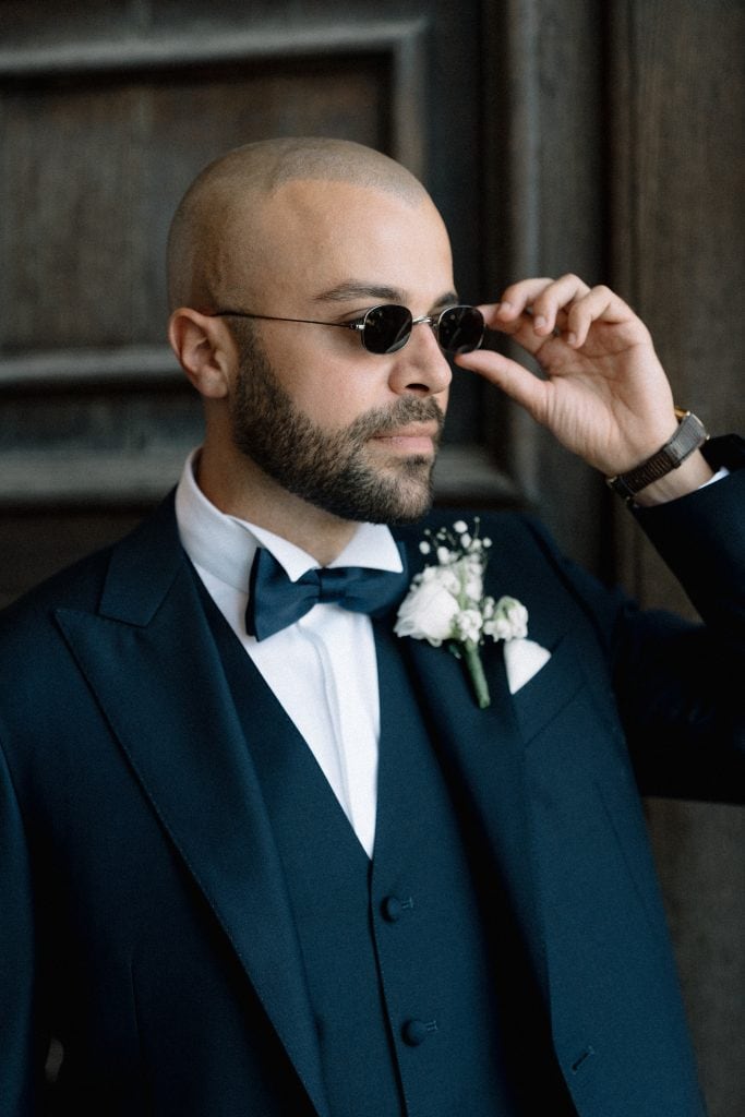 Sposo elegante con occhiali da sole, abito blu scuro e fiore bianco, in un contesto raffinato, pronto per il matrimonio di Alberto e Valeria.