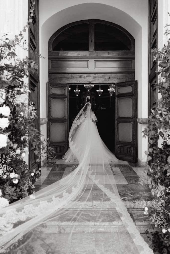 Sposa in abito da sposa con lungo velo che entra in una chiesa storica, circondata da fiori e decorazioni floreali, pronta per il matrimonio di Alberto e Valeria.