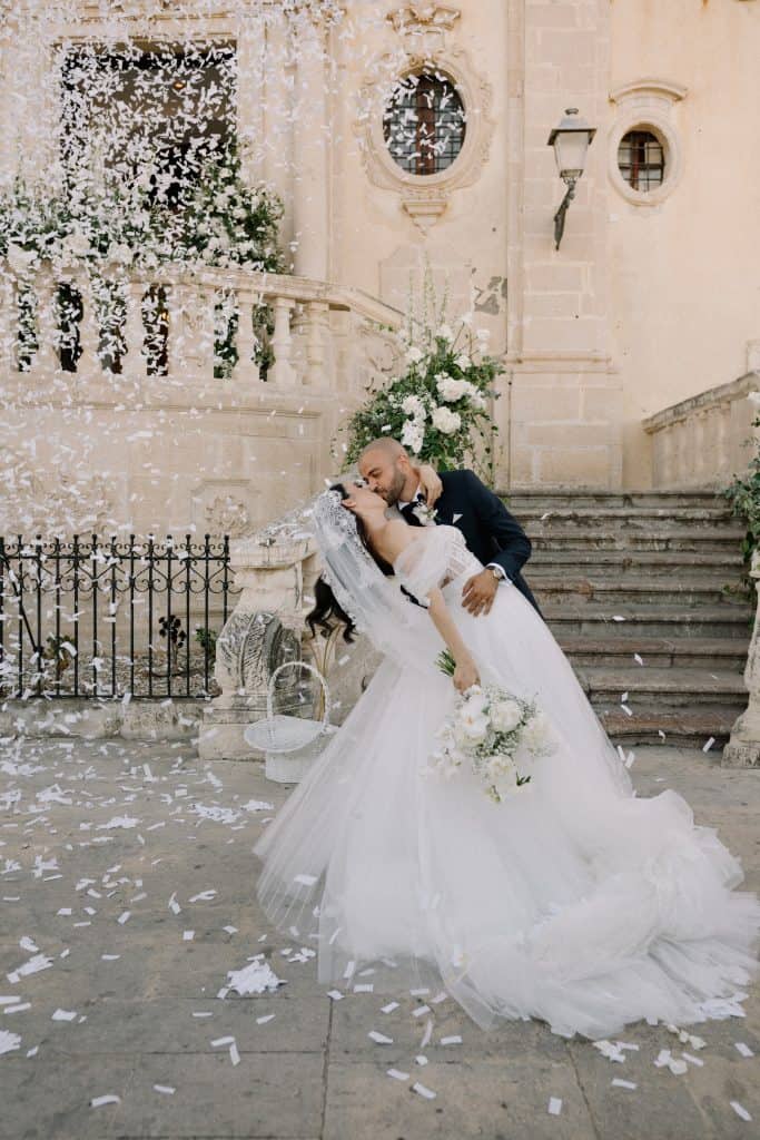 Alberto e Valeria si baciano durante il loro matrimonio, circondati da petali di fiori e decorazioni eleganti, in un suggestivo scenario storico.