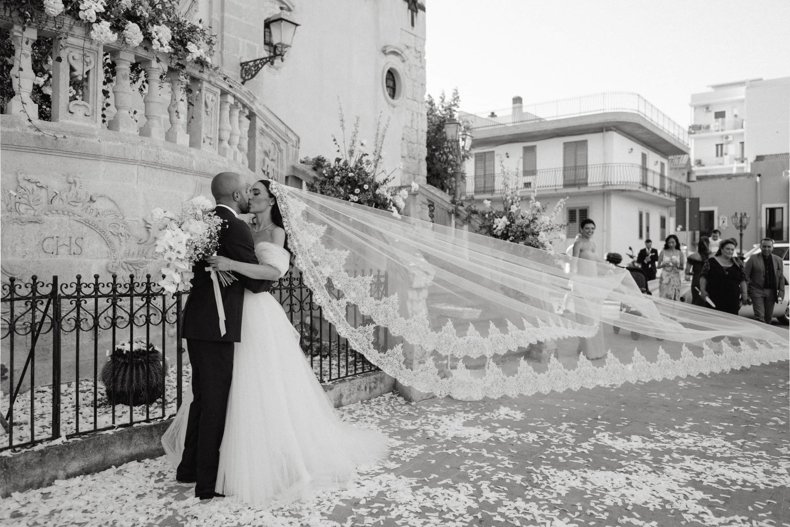 Házaspár, akik hosszú fátyollal csókolóznak az esküvőn, elegáns városi környezetben.