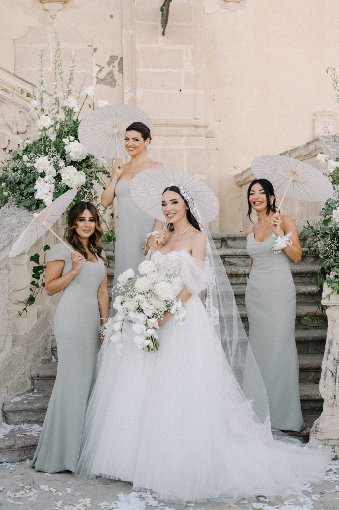 Sposa e damigelle con ombrelli bianchi durante un matrimonio elegante, decorazioni floreali e scenografia storica.