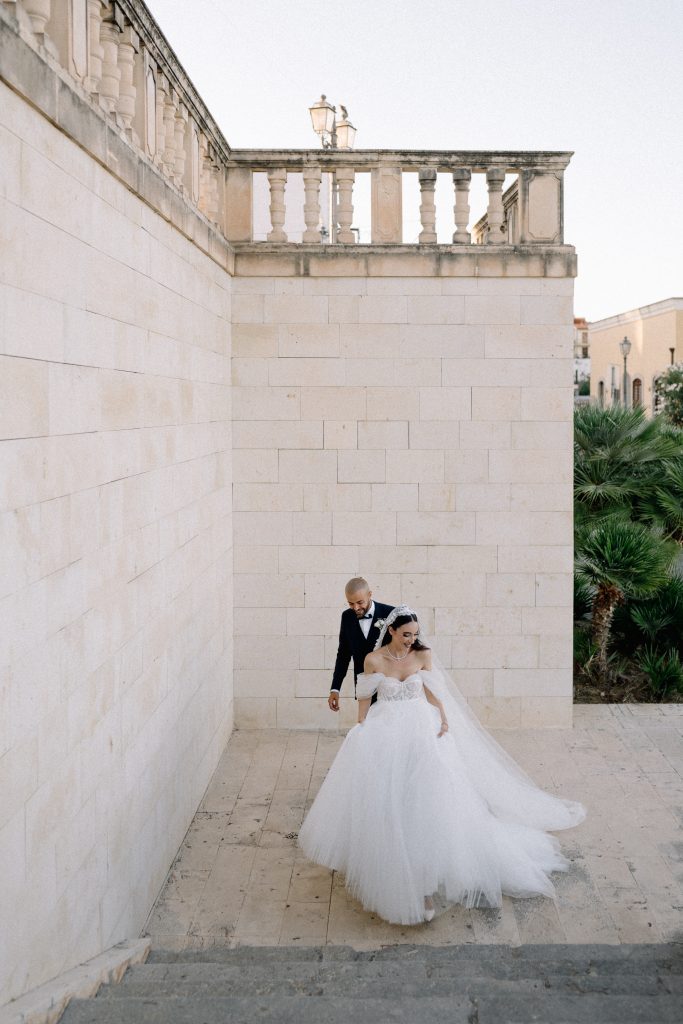 Alberto e Valeria durante il loro matrimonio, con la sposa in abito bianco e lo sposo in abito scuro, in un suggestivo scenario urbano con pareti in pietra e vegetazione sullo sfondo.