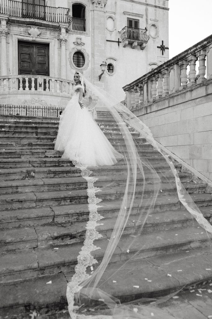 Sposa in abito da sposa con velo lungo e dettagli raffinati, posata sulla scalinata di un edificio storico, perfetta per un matrimonio romantico e sofisticato.