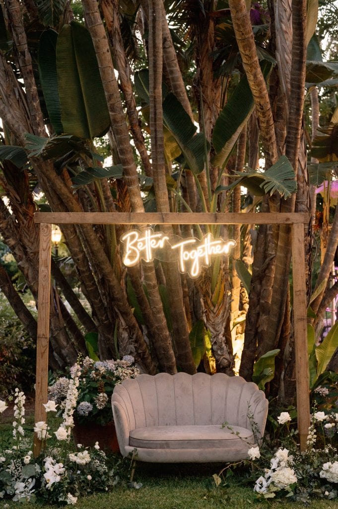 Neon "Better Together" illuminato su sfondo di piante tropicali, decorazioni floreali bianche e divano vintage, perfetto per un matrimonio romantico all'aperto.