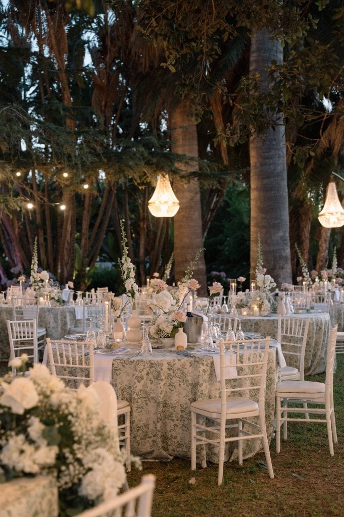 Tavoli eleganti per matrimonio all'aperto decorati con fiori bianchi e candele, ambientazione romantica tra gli alberi con luci sospese, perfetta per un matrimonio indimenticabile.