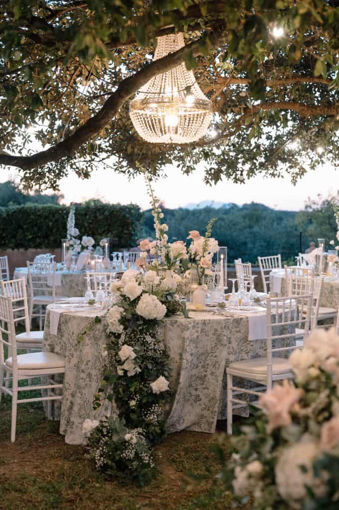 Decorazione floreale elegante per matrimonio all'aperto con tavoli e sedie bianche, sotto un grande albero con lampadario di cristallo, ideale per eventi romantici.