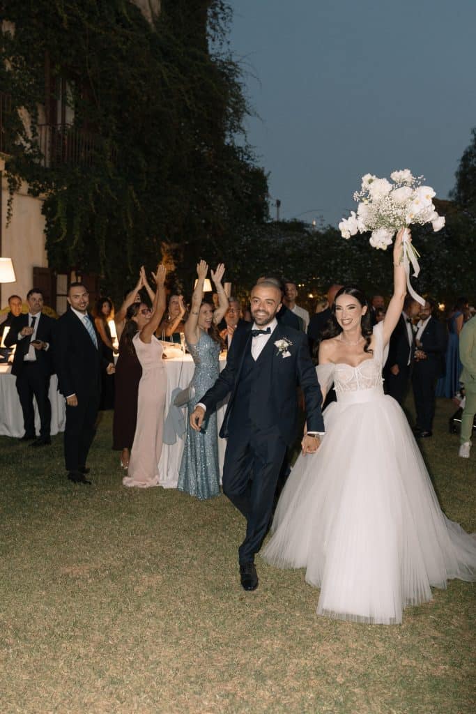 Alberto e Valeria camminano mano nella mano durante il loro matrimonio, circondati da amici e familiari in un giardino all'aperto, con decorazioni floreali e un'atmosfera di festa.