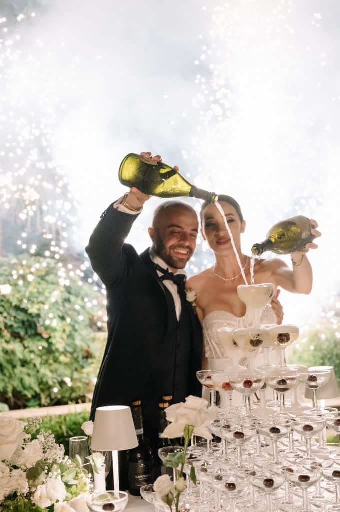 Coppia di sposi Alberto e Valeria brindano con champagne durante il ricevimento di nozze, decorato con fiori bianchi e un elegante tavolo con champagne.